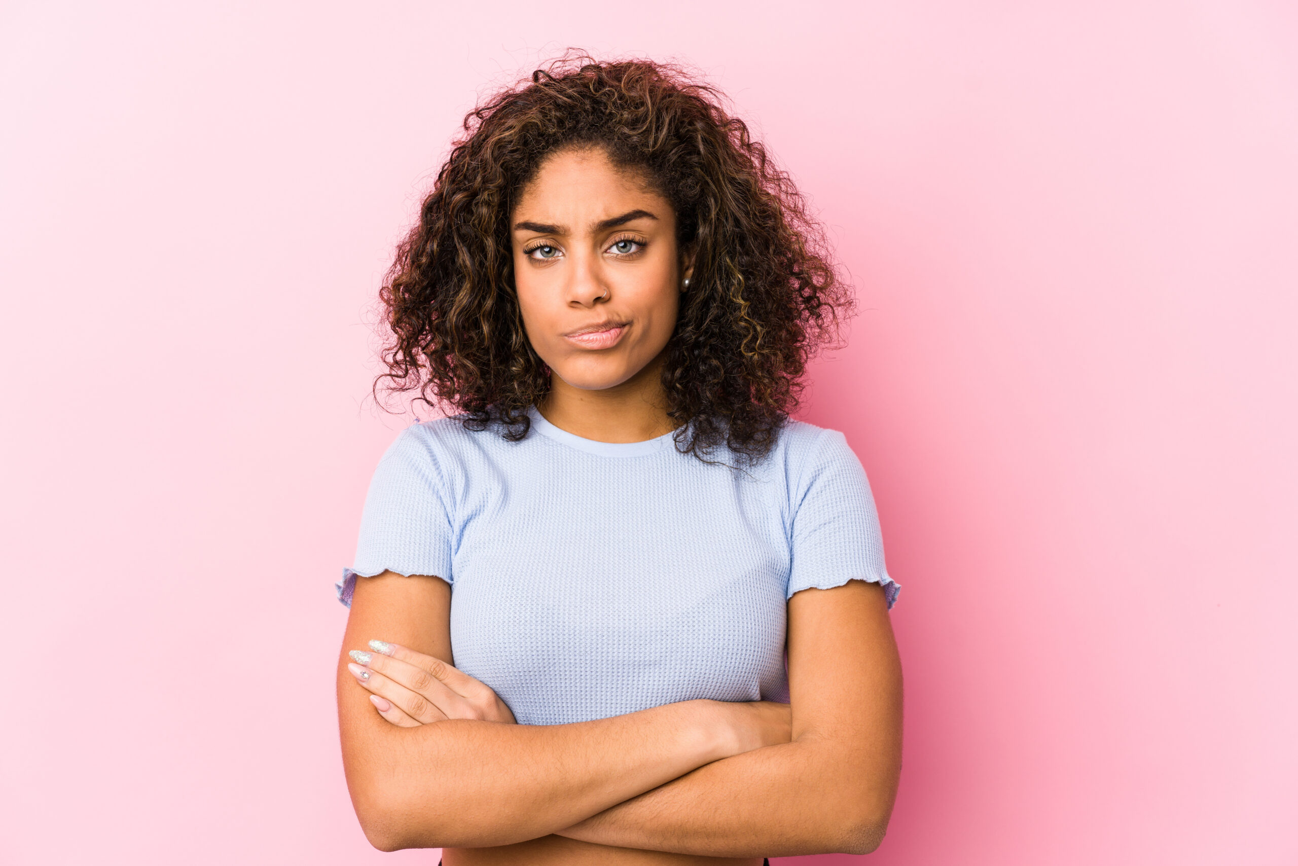 Young african american woman against a pink background frowning face in displeasure, keeps arms folded.