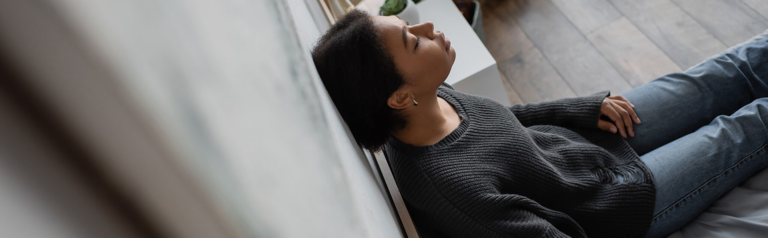 High angle view of depressed multiracial woman leaning on wall while sitting on bed at home, banner,stock image