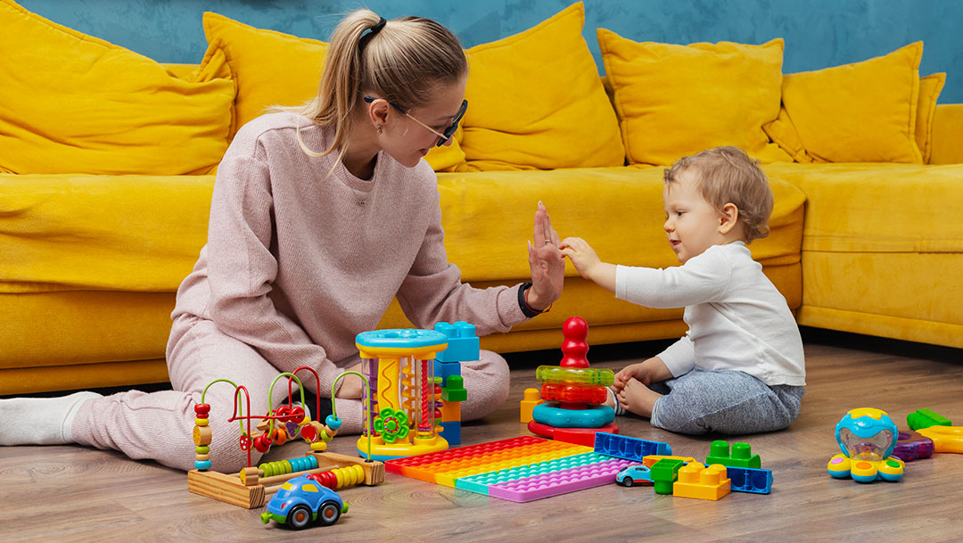 Assistante maternelle assise au sol avec un jeune enfant, jouant avec des jouets d’éveil dans un espace intérieur aménagé.