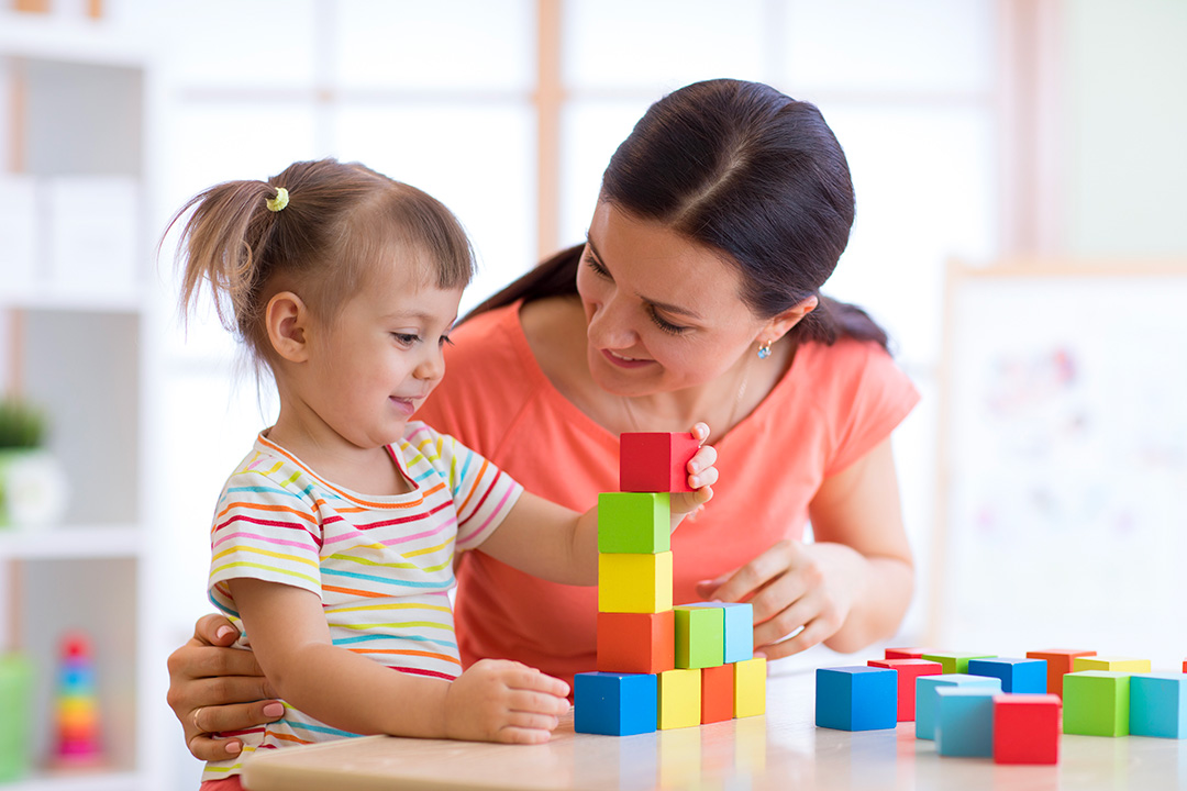 Assistante maternelle accompagnant un jeune enfant qui empile des cubes colorés sur une table, dans un espace intérieur lumineux.